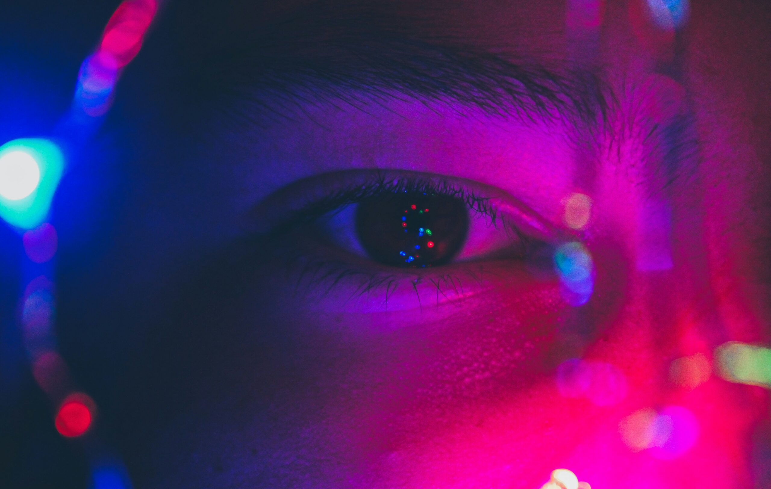 A close-up of a person's eye illuminated by colorful neon lights, with reflections and blurred light spots surrounding the face—capturing how plants see light differently in their own vibrant world.