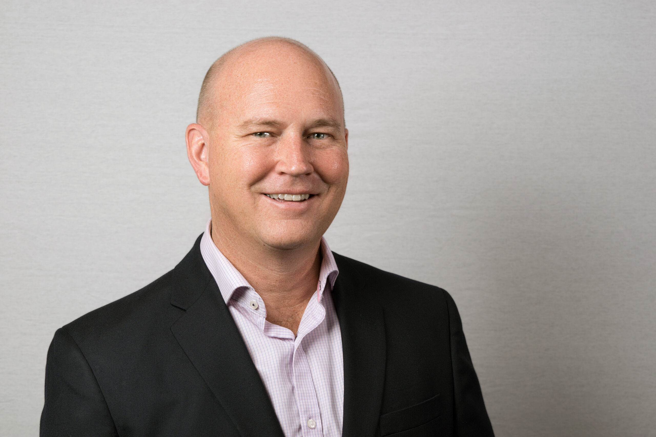 A man with a shaved head, wearing a black suit and pink shirt, smiles confidently against a plain gray backdrop, embodying the sleekness of new technology.