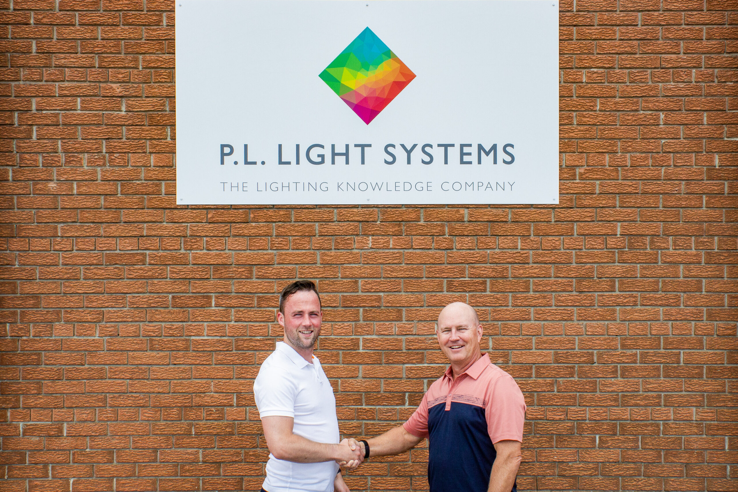 Two men shake hands in front of a brick wall with a P.L. Light Systems sign, featuring a colorful geometric logo above the company name—symbolizing a new strategic partnership between the parties.